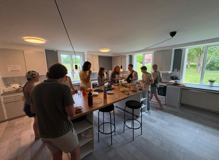 nine students stand having conversations around a wooden kitchen island with various dishes and drink options