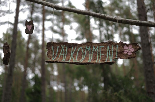 a piece of bark with Valkommen written in blue writing on it hangs with twine from a branch with forest trees in the background