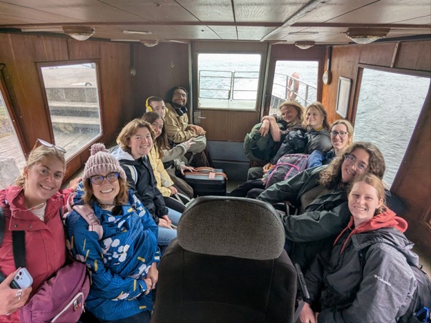 11 people sit on 2 rows within a small boat with the wood of the dock and water visible out of windows to either side