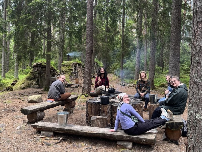 Cooking over the fire with sleeping huts in the background in a forest.
