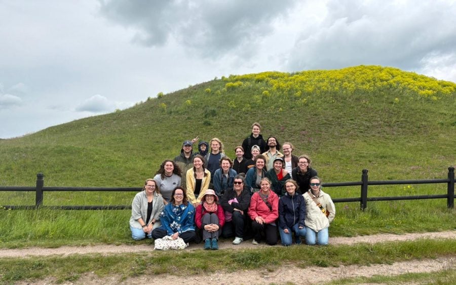 a group of smiling students and teachers sitting in front of a large grassy mound with a wooden fence right behind them