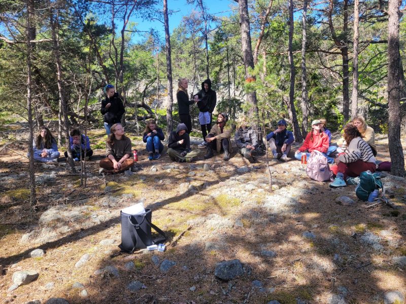 a group of students are seated on the ground in a pine forest while a women is walking around handing out something to them