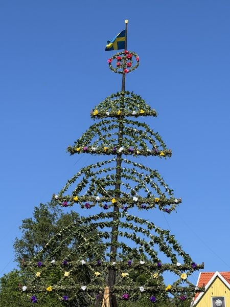The midsummer maypole from Arlhoma is shown against a blue sky, with 4 rainbow shaped arches, a circle at the top, topped with the Swedish flag. 