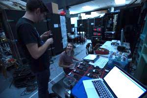 Samantha Palumbo seated at a sound board in an equipment room talking with another technician